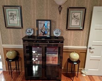 Vintage hand-painted black lacquer lighted display cabinet, with two glass shelves, pair of mahogany camp tables, with pair of crackle glazed yellow Asian jars, Asian doll in glass display case, flanked by a pair of Asian porcelain plates and pair of nicely framed/matted botanical prints.