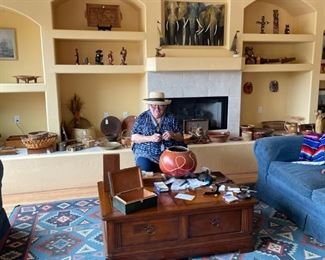 Barbara, hard at work and surrounded by a wonderful coffee table, Dhurrie rug, two blue love seats, and a back wall filled with Tribal arts featuring gourds, baskets, tribal carvings and more. There's also a Diana Maderas serigraph of elephants from her time spent in Africa.