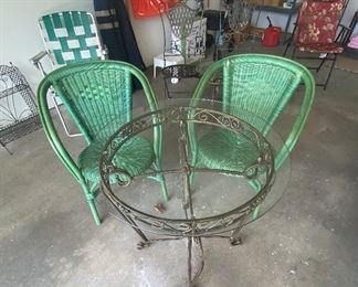 Large metal table with glass-top. Two green wicker chairs.