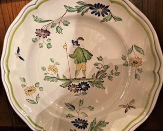 "Moustiers by Longchamp" - fine china dinnerware from France which features images of a man with a feathered cap and walking stick surrounded by floral designs in maroon, dark blue, and yellow