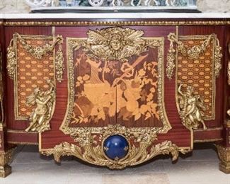 A Louis XIV Style Commode in the Manner of Jean Louis Riesener. Probably Twentieth Century. No Marks Evident.

 The commode in the Louis XIV style and of demilune form having a demilune top with conforming white marble slab over the case, the edge with applied brass egg and art molding over multiple marquetry inlaid panels bracketed by large yellow metal figures, floral and foliate swags and putti; the center with two cabinet doors bracketed by applied yellow metal cornucopia and mask with sunburst rays and applied cerulean blue orb, raised on tapered feet with applied yellow metal acanthus leaf decoration. Dimensions: Height 39 x width 98 x depth 29 inches.  $45,000.00

