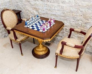 A Pair of Louis XVI Style Fauteuils. Circa Last Quarter of the Twentieth Century. No Marks Evident.
The pair of fauteuils in the Louis Seize style with embellished yellow metal accents in the form of a wreath and ribbon finial at crest rail and applied metal accents to the skirt together with a Tiche burl walnut and gilt wood chess table having inset porcelain chess board, Tiche porcelain chess pieces and raised on a vasiform gilt wood standard ending on a round plinth.

Dimensions: Chairs Height 40 ½ x width 23 x depth 18 inches. Table Height 30 ½ x width 32 x depth 32 inches.   For the suite $8,000.00
