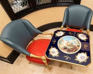 6An Empire Style Table and Leather Upholstered Chairs. Tiche. Italy.  
TheTiche table in the French Empire style having an enameled top with inset porcelain placques, the center medallion with Tiche mark and depicting a classical scene of putti surrounded by four medallions with figures in 18th century attire, raised on gilt medal winged figured joined by a medial shelf with white bisque porcelain sculpture of two female nudes ending on scrolled toes. The two chairs having blue and red upholstery with gilt wood frames.
Dimensions: Table Height 31 x width 27 x depth 27 inches. Chairs Height 41 ½ x width 26 ½ x depth 24 inches.    For the suite $10,000.00
