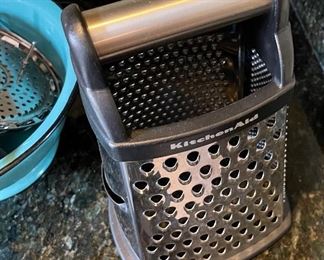 ot 8341. $38.00. Kitchen collection: Kitchen Aid Gourmet 4 sided box grater; turquoise 9" metal colander -made in Poland; red, Good Cook apple cutter; steamer basket' vegetable peeler; serving spoon and fork and a small scale.