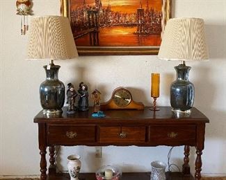 Beautiful wooden entrance table with an assortment of decorative items on top of it. Two large lamps with shades frame a lovely orange sunset canvas, with an owl wall clock beside the painting.