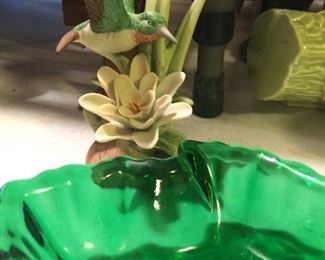 A delicate porcelain bisque hummingbird among a green dish and ceramic planter. 