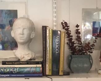 Book shelf with various art books, 2 Original Art Pieces, Antique Brass Candlestick, Art Glas Ball Bottle Vase, Bust Sculpture by Jane Paden, Vintage Roseville Vase, Antique MacBeth Figurineand a large Brass Pan,