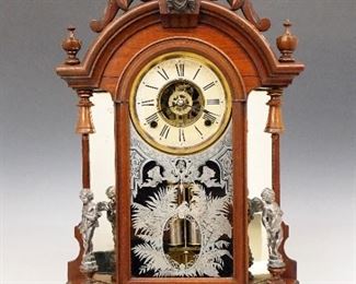Ansonia Triumph Model Shelf Clock - A late 19th century Ansonia "Triumph" model shelf clock.  8-day time, strike and alarm movement with a painted metal dial and Roman numerals.  Carved Walnut case with shaped crest, applied Liberty Mask and turned finials over a single arched door with stenciled glass, flanked by mirrored sides with cast Cherub figures, on a rounded molded base with applied metal decoration.  Paper label 50% intact.  Old finish with wear, some shrinkage, dial wear and discoloration, running when cataloged.  24" high. 