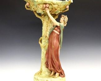 Lot 25: A turn of the century Royal Dux porcelain compote.  Depicts Eve picking the forbidden apple with multicolor glaze decoration and Gilded detail.  Applied lozenge mark with impressed numerals.  Some surface wear, wear to Gilding, a few tiny edge flakes, shallow chip to base.  19 3/4" high.  ESTIMATE $400-600
