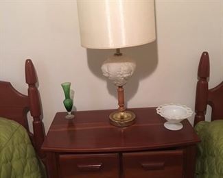 cherry bedside table sits between two matching twin beds , table has mid century lamp with a piece of milk glass and green vase 