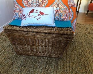 Large woven trunk with handles; jute rug with orange/teal accents.