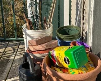 Plastic planters and watering can, metal yard stakes/holders, open bag of potting soil and open canister of plant food.