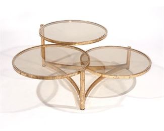 Lot 41 | MILO BAUGHMAN THREE TIERED SWIVEL TABLE | Tri-level brass and glass coffee / low table, having smoky glass on a polished brass frame; h. 17 w. 34 in.