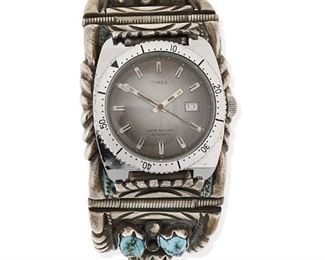 6124
A Ray James Jr. Navajo Watch Band
Fourth-Quarter 20th Century, Diné
Stamped: R. James Jr. [for Ray James, Jr.]
A heavy cuff with nine turquoise nuggets set in large tooth bezels, with incised edges and spring clasp closure, centering a Timex watch
7" total inner C (closed) x 1.25" H
250.0 grams
Estimate: $400 - $600