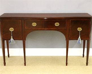 A Baker Hepplewhite style sideboard.  Mahogany construction with Satinwood banding and Ebonized inlays.  Shaped top with molded edge detail over a central silverware drawer with flanking concave doors, on tall tapered legs ending in spade feet.  Very slight wear.  20 1/2 x 68 1/2 x 38" high overall.  ESTIMATE $800-1,200