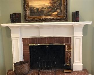 Living Room:  A landscape painting in an ornate frame hangs above the mantel which displays five faux leather storage books.  On the hearth are two wood log holders and a set of brass fireplace tools.