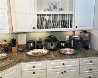 Kitchen Counter:  Displayed are a copper chafing dish; CUISINE DE FRANCE knife set block; PRESTO fryer; WEST BEND air popper; INSTANT POT; electric OSTER fondue pot; BREVILLE juice fountain; WARING Pro-Ice Crusher; a large turkey platter; and several silver chargers.  Above those items are a three piece porcelain HUTSCHENREUTHER coffee set and several decorative plates.  