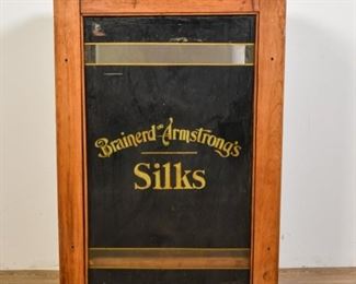 334	Brainerd and Armstrong's Silks Mercantile Cabinet	Brainerd and Armstrong's Silks Mercantile cabinet. American, Late 19th Century. Wood with brass hinges, 4 shelves, and black reverse painted glass front. Glass is missing from other side of cabinet. Heavily worn on top and sides. 36" H x 24" L x 11" D
