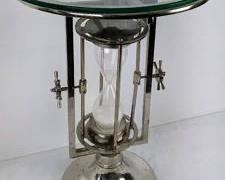 This eye-catching novelty Dulin Furniture turning sand-timer table actually works! The circular glass top sets on a fixed frame, while the white sand-filled glass hourglass mechanism flips beneath. The chrome finish shows some tarnish marks, mostly on the base. Small ship's-wheel turning handles rotate the sand-filled hourglass (which was not timed). The top glass of this table measures 17* wide. It stands 24" high with an 11" base. 