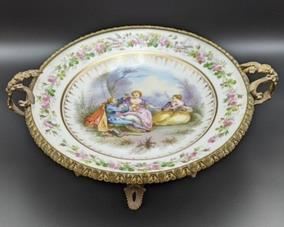 Sevres Painted  Porcelain Dish with Ornate Stand. Lovely painted scene dish with gold detailing rests atop a decorative footed brass stand with ornate details. The stand needs some repair and the plate sits crooked as seen in the photos. Measures 14” in diameter and 5.5” high