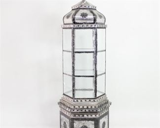 Lot 56: Punched Tin Hexagonal Embossed Metal Glass Display Case. This glass display vitrine recalls Islamic architecture in style, with elaborate embossed metal and other decorations, including pierced carvings and fretwork in alternating geometric and floral motifs. Upper section is six rectangular clear glasses framed with silvered metal and a door for access to internal compartments. Three shelves, two made of glass and one mirrored at the bottom. Showcase cabinet is wired for illumination from above via a top compartment behind the lidded cover with ball finial. Removable hexagonal base also has a storage compartment. A unique display piece for your home or retail showroom.

Dimensions are 67.75 in h x 22.5 in w x 22.5 in d. Condition is fair, with dents and oxidation throughout, and two cracks in glass on one side. Inline switch on cord has not been tested. Please call or email for a detailed condition report.