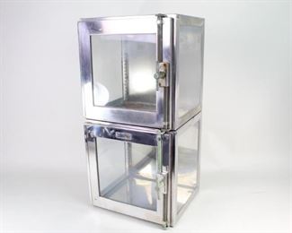Lot 337: Pair of Metal Stacking Glass-front Medical Boxes Cabinet. These vintage industrial glass-fronted cubes have brackets on the inside to allow for interior shelves, and round indents in each corner that keep them in place when stacked. Left-hinged doors with an attractive metal turn handle. Small enough to fit on a deep tabletop or countertop. One unit has a cutout in back where electrical for lighting etc. could be brought in. Probably once used for medical chemicals, they could display your sundries in a contemporary kitchen or retail setting.

Dimensions are 12 in x h 12 in w x 12 in d. Condition is good, with surface wear throughout, small rust spots, wear to inside including marks at bottom from previous liquid containers, and rubber gaskets that are cracked but still intact. Please call or email for a detailed condition report.