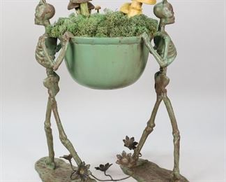 110	Skeleton Metal Plant Stand	"Skeleton metal plant stand, made of metal, with 2 skeletons holding a bowl of fake greenery and mushrooms. Wear and losses to patina.
16 1/2"" H x 11 1/2"" L"
