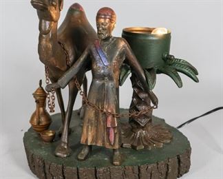 123	Painted Metal Orientalist Lamp	Painted metal orientalist lamp. Metal man and camel with palm tree lamp and small metal ewer and cup, mounted on wood base. 11 3/4" H x 11 1/4" wood base diameter. Later painting to the man and camel figures. Wear to paint back of camel's head. Pitting to the man's body.
