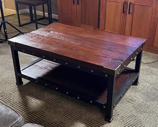 INDUSTRIAL COFFEE TABLE | Low table, having a dark wood board top and lower shelf with brass supports and tacks on a black metal block leg frame; h. 20 x 46 x 30 in.