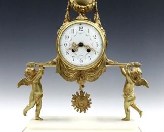 A late 19th century French mantle clock by Adolphe Mougin, Paris.  8-day time and strike movement with Apollo mask pendulum and convex porcelain dial with Arabic numerals and fancy filigree hands, serial #5399. Gilded Bronze case with an urn finial with foliate garlands above two Cherubs holding the movement aloft on a White Marble base with gilded frieze and beaded feet.  Slight wear,  running when cataloged.  17 3/4" high.  ESTIMATE $1,000-1,500  