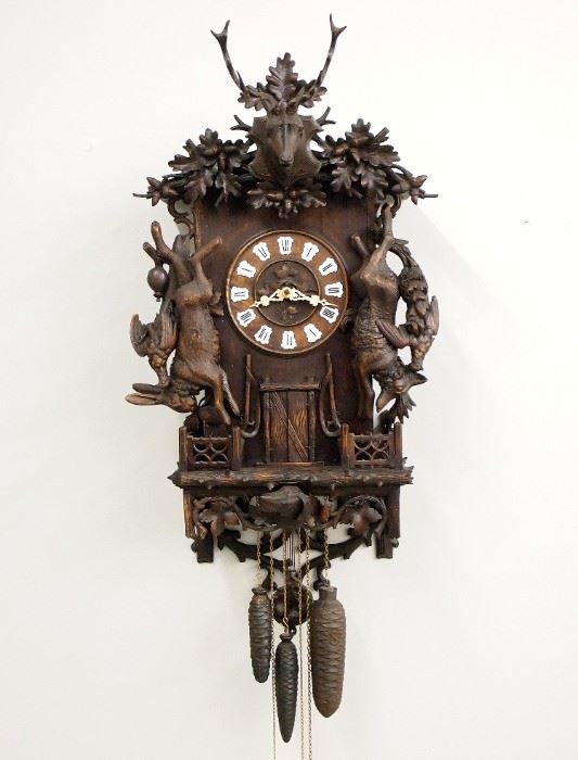 A turn of the century Emilian Wehrle 5 Horn Trumpeter Black Forest wall clock.  Brass 30-hour three train movement with arched Brass plates features a carved wooden dial with Oak leaves at the center and porcelain hour markers with Roman numerals, announces the hour with a melody played on five horns, counts the hour on a coiled gong, carved Oakleaf pendulum bob.  Carved Walnut case with a pediment top features a carved Stag's head with an Oakleaf crest, surmounting hanging game and long rifles flanking a door which opens to reveal a German horn player automaton, with a scrolled lattice fence and lower Oakleaf skirt with dog's head and scrolled pack panel.  Partial paper label on bellows.  Old dark finish with some wear, repair to rabbit's ear, repair to 12:00 marker, movement restored and running when cataloged.  44" high plus weight drop.  ESTIMATE $4,000-6,000