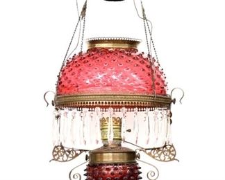 A turn of the century kerosene hanging lamp.  Cranberry Glass hobnail shade with matching font, in an embossed Brass frame with one row of faceted glass prisms.  Electrified, some wear and minor damage, soldered repair at burner.  Approx. 35" high.  ESTIMATE $600-800