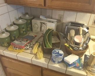 Vintage 4 piece canister set green metal with flowers, Salton bun warmer electric, Blue Holland Tin, 5 Pot Holders, Knitted Corn on Cob, Black and Decker 2 Slice Toaster, Meat Slicer. Various Kitchen Utensils in Basket.