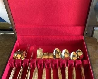 Japanese gold colored flatware set in wood case, includes butter knives, serving ladle, serving fork, spread knife, and multiple sizes of forks and spoons. Case has red felt-like material inside, with scratches and scuffs to surface of case.