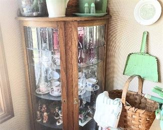 Corner curio cabinet filled with Hummels, cup and saucer sets and cranberry glass. The pitcher on top is Longaberger.