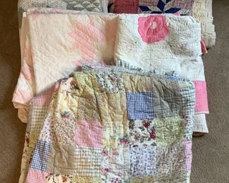 Includes (5) large quilts, (4) appear to be homemade. One patchwork style quilted bedspread in pastel florals, (1) white with pink flowers, (1) white with pink squares, (1) white green tree pattern and (1) pink with multi-colored pinwheel and white border. Some may have tears, worn areas or stains.
