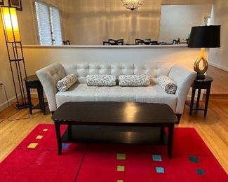 Chateaux D'ax white leather sofa with black rectangular rounded corner coffee table & two matching 1 drawer end tables atop a red geometric carpet. 1 gold table lamp & 1 glass shaded floor lamp