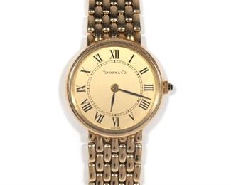 Lot 225  |  LADY'S TIFFANY & CO. 14K GOLD WRIST WATCH  |  Gold face with Roman numeral markers, sapphire crown, on a 14k gold tread-link bracelet; 7.5 in., 38.6g