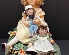 R. John Wright Marcella, A Raggedy Ann Story Doll Set. A wonderful nostalgic set, a true must for any doll collector!
Includes Marcella holding small a Raggedy Andy doll with Raggedy Ann at her feet while sitting on a floral decorative green/grass like stand. She comes with a box and certificate. She is number 13/100. There is also a box for the green stand she is sitting on.
Name: Marcella, A Raggedy Ann Story™
Description: 17" tall, Marcella constructed of felt with a molded face with hand painted features and ball-jointed knees. Dress of custom-dyed cotton gauze with hand-cobbled leather shoes and felt and silk cloche-style hat. Custom-made display base made of wood and molded felt covered with handmade felt flowers and leaves. Includes 6" Raggedy Ann™ and Raggedy Andy™ also made of felt with hand painted features. Diameter of the base: 14"; overall height: 16".