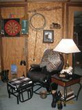 Black Tubular Metal Glider Rocker with Matching Foot Stool, Black Metal Frame Lamp Table with Glass top and Wood Trims. Back Ground Marlboro Dart Board, Lasso, etc. Pair of Kangaroo Boots in fore ground.