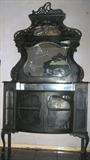 Victorian Etarge're on cabinet base raised on delicate pad feet Queen Anne legs, ornately carved Carved scroll shelf supports. Cabinet has a bow front with curved glass double doors. Top shelf has original brocade faric lining. Bevel crest shape center mirror and a smaller bevel mirror just below the crown. All appears to have original finish, mirrors & glass. The overall measurements are 89"H x 48"W x 16"D