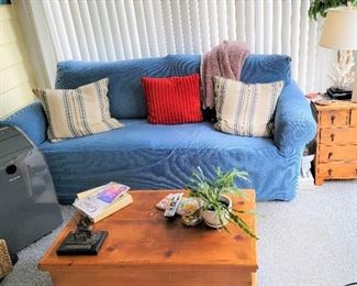 Several items in this photo include a Blue Denim Sleeper Sofa (with a cover over it) a portable Air Condition Unit, A very nice wooden Coffee table that opens at the top for storage, and a cute four drawer side table.