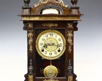 An early 20th century Hong Kong-American Bell top shelf clock (Hamburg-American Clock Co. for Chinese Distribution).  8-day time and strike movement with a painted metal dial, Arabic numerals and an embossed Brass center and bezel.  Mixed wood case with applied Brass decoration and shaped crest over a single long door and bracket base.  Paper label 80% intact, additional label on back.  Original dark finish with some wear, movement dirty, runs momentarily.  23 1/4" high overall.  ESTIMATE $300-400
