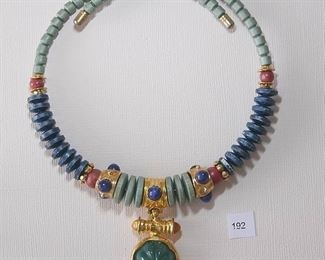 Francesca Romana Choker style necklace measures approximately 18” in length and is strung on a tight metal wire.  Half of the choker is light blue opaque beads. Part of the choker is a darker, larger opaque bead.  The two colors separated in the side center by a round reddish bead and gold tone Rondells.  The center of the choker is a mix of light blue flat beads, gold tone sections with a dark blue opaque cabochon and red opaque beads. A center drop is held by a textured gold tone bar with yellowish round cabochons.  The bar holds a round fluted dark green opaque cabochon in a gold tone bezel.  No clasp as the tight metal wire holds the necklace in place.