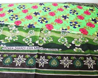 Two table coverings/cloth fragments, one African with Swahili Proverb “POKEA ZAWADI KWA MIKONO MIWILI” which translates as “RECEIVE GIFTS WITH OPEN ARMS”, pieced together to form a 56” by 41” table cover, marked LESSO PRINTED KHANGA TANZANIA DES NO 9150, and one Japanese 12” by 122”, marked PRINTED ACRYLIC NIAGARA.