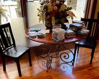 Round solid cherry topped dining table with fancy ironwork base and two black side chairs.