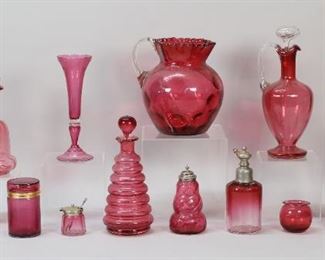 331	13 Pieces Cranberry Glass	"13 pieces cranberry glass. 2 decanters with handles, vase with etched grape and leaf decoration, bottle (no stopper), ""beehive"" decanter, small jug, 2 muffineers with silverplate tops, cabinet vase, condiment jar with silverplate lid and silver spoon, cylindrical box with hinged lid, coinspot pitcher, atomizer (missing bulb). Silver spoon 5.1 grams. Pitcher 7 5/8""H, largest decanter 10 5/8""H including stopper.

Roughness to end of stopper on smaller handled decanter, chips to end of stopper on beehive decanter, internal crack to handle of jug, chip to rim of cabinet vase, smaller muffineer chipped on rim below collar."
