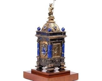 LAPIS LAZULI & SILVER MOUNTED CLOCK TOWER | In gothic style, having a domed top with a knight-form finial holding an enameled shield, silver base with lion motif, tower sides with lapis cartouches, engraved clockface with Arabic numerals behind a beveled glass door; the silver hallmarked on one side with "VN" in conforming reserve / "HJ" in a rectangle, and with an indistinct hallmark (possibly Scottish), mounted on a stepped wood platform
h. 14-3/4 w. 4-1/2 d. 4-1/2 in. (clock only)
overall: h. 16-1/2 x 7-3/4 in. (on platform)