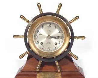 CHELSEA MARINER SHIP BELL CLOCK | CHELSEA "Mariner" Helm Ships shelf or wall clock. A Chelsea Clock Co. (Boston) Mariner model ship's clock with Ships Bell movement. Brass and bronze ships bell clock in form of ships wheel. Silvered dial with Arabic numerals and marked Chelsea Ship's Bell. Eight-day lever escapement movement with ship's bell strike. Serial number 583973. Mahogany base. Dedication plaque has a date of December 16th, 1953.