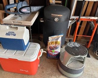 Garage:  A small tray table on iron base; a RYOBI 18 volt cordless chain saw with blade cover;  and coolers are to the left of a BRINKMANN Smoke 'N Grill; hand cart; and vintage stool. 